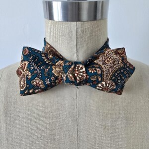 May include: A teal paisley bow tie with brown and tan floral accents. The bow tie is displayed on a mannequin. The fabric has a detailed pattern, adding a touch of elegance. This accessory is suitable for formal events.