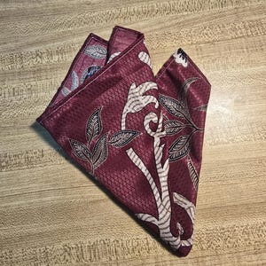 May include: A folded burgundy pocket square with a detailed floral and abstract design. The fabric features a repeating pattern of small diamonds and is accented with white and gray botanical elements. The pocket square is displayed on a light brown wooden surface.