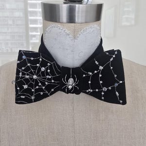 以下が含まれることがあります： 中央に銀色の刺繍が施されたクモ、両サイドに銀色の糸で描かれたクモの巣があしらわれた黒いベルベットの蝶ネクタイ。クモの巣の糸には、露のしずくを模した小さな透明なビーズが飾られています。