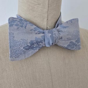 May include: A light blue bow tie with a white and light blue patterned design. The pattern features a repeating design of trees and a small boat.