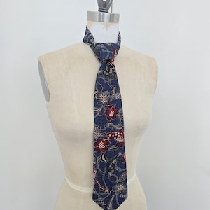 Puede incluir: Una corbata azul marino con un estampado floral en rojo, gris y blanco. La corbata se exhibe en un maniquí. El diseño floral incluye flores y hojas estilizadas. La corbata está anudada al cuello.