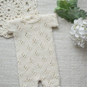 May include: Ivory-colored knitted baby romper with short sleeves and openwork diamond patterns. The romper has a round neckline and scalloped edges. A crocheted doily and white flowers are in the background.