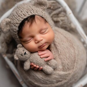 Puede incluir: Un bebé recién nacido con un gorro de oso de peluche de punto marrón y envuelto en una manta de punto marrón, sosteniendo un oso de peluche de punto marrón.
