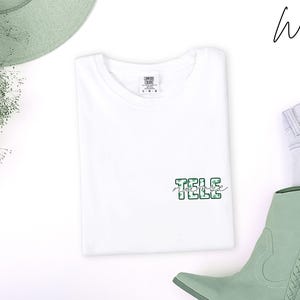May include: A white t-shirt with the text "TELE Nurse" in a green and white camouflage pattern. The shirt is laid flat with a pair of light green cowboy boots, light blue jeans, and a green hat.