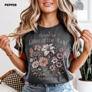 以下が含まれることがあります： 「Consider the Lillies of the Field」と「Matthew 6:28」の文字と花柄がプリントされた、濃いグレーのTシャツ。 白、ピンク、ピーチ色の花がアレンジされています。 左上隅には「PEPPER」の文字があります。