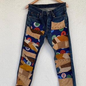 May include: Dark wash denim jeans with patchwork details. The jeans feature various brown, tan, and blue fabric patches with decorative stitching. The patches include a blue fabric with a colorful circular pattern. The jeans are displayed on a wooden hanger.