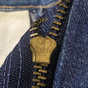 May include: Close-up of a brass zipper on blue denim fabric. The zipper is partially closed, revealing the interlocking teeth and the brass pull tab. The denim has a textured appearance with visible stitching.