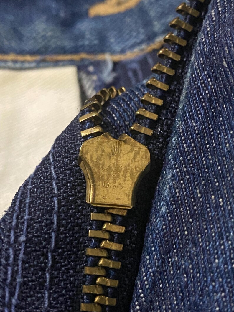 May include: Close-up of a brass zipper on blue denim fabric. The zipper is partially closed, revealing the interlocking teeth and the brass pull tab. The denim has a textured appearance with visible stitching.