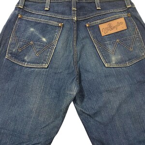 May include: Back view of blue denim Wrangler jeans. The jeans have two back pockets with the Wrangler logo stitched on the right pocket. The jeans have a classic five-pocket design with a button and zipper closure.