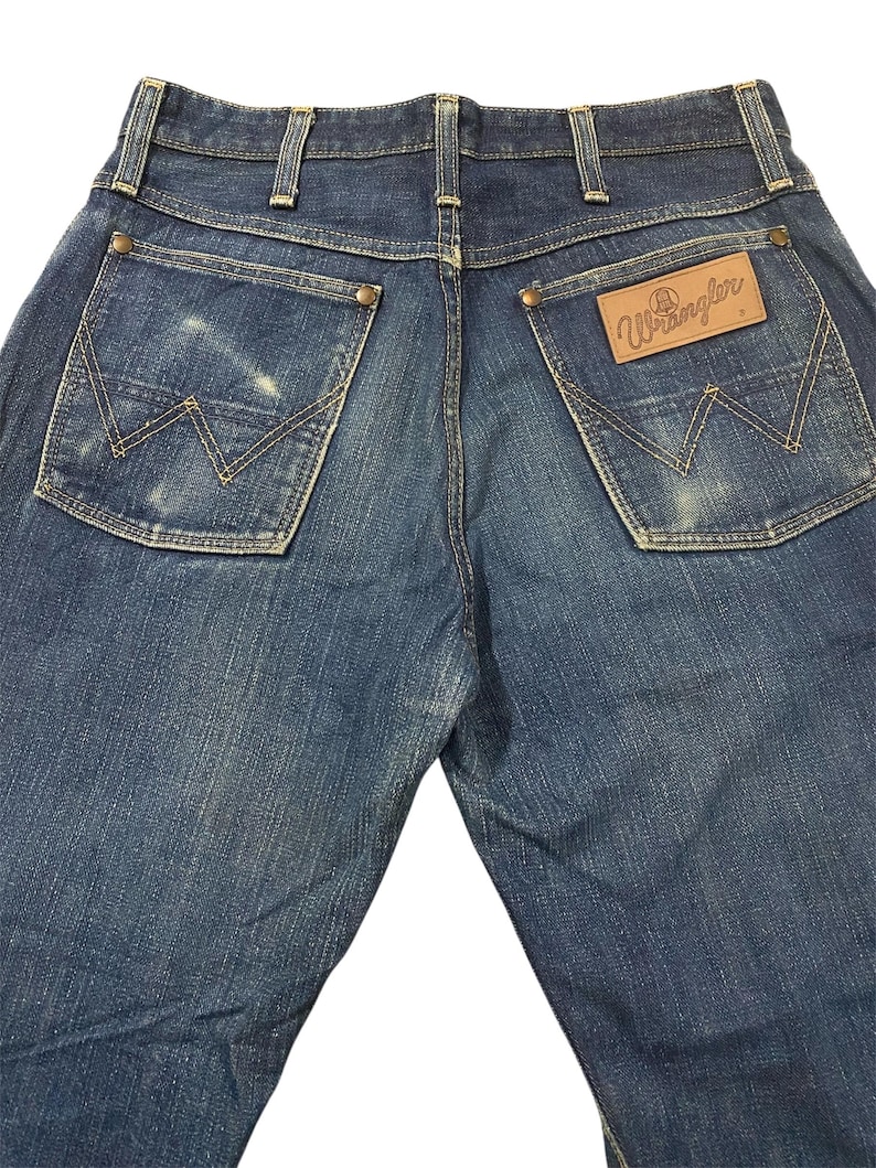 May include: Back view of blue denim Wrangler jeans. The jeans have two back pockets with the Wrangler logo stitched on the right pocket. The jeans have a classic five-pocket design with a button and zipper closure.
