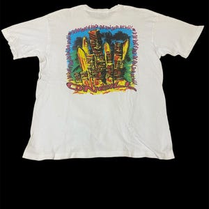 May include: White t-shirt with a colourful graphic on the back featuring tiki-style figures, surfboards, and the brand name "O'Neill." The design includes text around the border. The shirt has short sleeves and a crew neck.