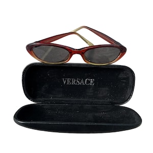Pu&ograve; includere: Occhiali da sole ovali rosso-marrone con custodia nera. Gli occhiali hanno una montatura sottile e lenti scure. La custodia &egrave; aperta e presenta la scritta "VERSACE" stampata in bianco.