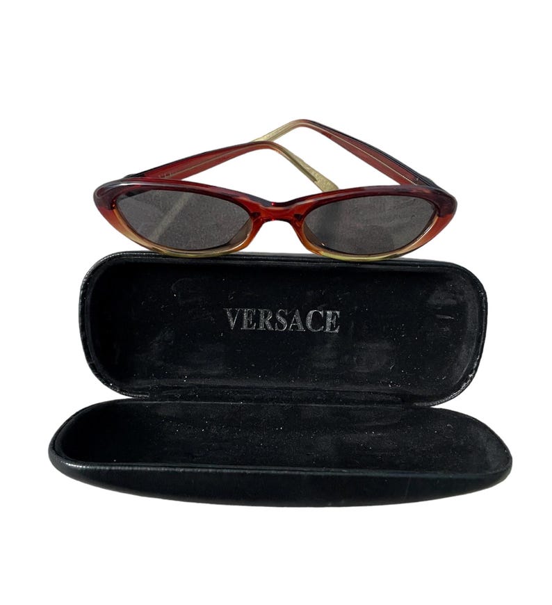 Pu&ograve; includere: Occhiali da sole ovali rosso-marrone con custodia nera. Gli occhiali hanno una montatura sottile e lenti scure. La custodia &egrave; aperta e presenta la scritta "VERSACE" stampata in bianco.