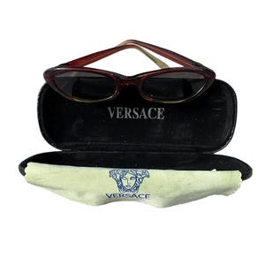 Pu&ograve; includere: Occhiali da sole marroni a occhi di gatto con custodia nera. La custodia &egrave; aperta, rivelando un interno nero e la scritta "VERSACE" in bianco. &Egrave; visibile anche un panno per la pulizia verde chiaro con il logo Versace.