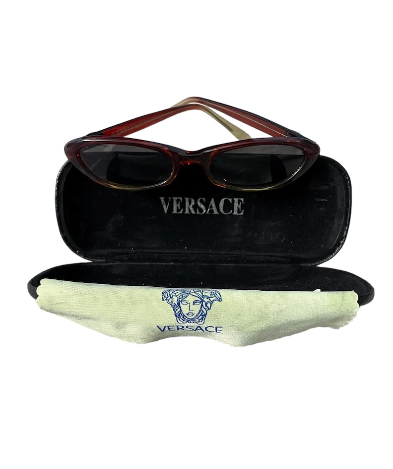 Pu&ograve; includere: Occhiali da sole marroni a occhi di gatto con custodia nera. La custodia &egrave; aperta, rivelando un interno nero e la scritta "VERSACE" in bianco. &Egrave; visibile anche un panno per la pulizia verde chiaro con il logo Versace.