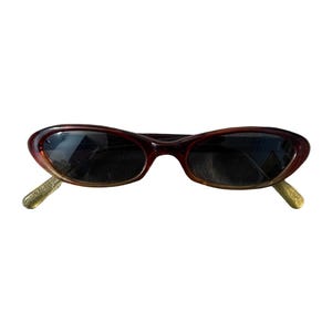 Pu&ograve; includere: Occhiali da sole ovali marroni con lenti scure. La montatura passa da un marrone scuro a un marrone pi&ugrave; chiaro. Le aste sono di colore oro chiaro. Questi occhiali da sole sono un accessorio elegante.