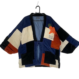 May include: A quilted patchwork jacket featuring a color-blocked design in navy blue, black, cream, and rust orange. The garment includes a black collar, two front pockets, and an embroidered carrot graphic with the text BRITISH RABBIT.
