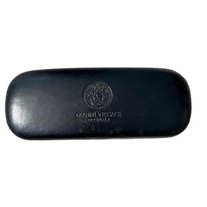 Pu&ograve; includere: Astuccio rettangolare nero per occhiali con la scritta in rilievo "GIANNI VERSACE OCCHIALI" e un emblema circolare. L'emblema presenta un disegno a testa di Medusa. L'astuccio &egrave; realizzato in un materiale liscio, possibilmente simile alla pelle.