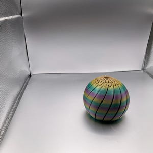 May include: A colorful, spherical decorative object with a striped pattern in shades of blue, green, yellow, and purple. The top section has a carved, wooden-like design. The object is sitting on a white surface.