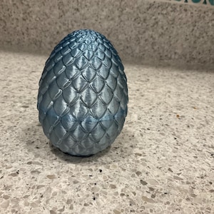 May include: A blue decorative egg with a textured, scaled surface. The egg is a light blue color and has a rounded shape, resembling a dragon egg. The egg is a decorative item.