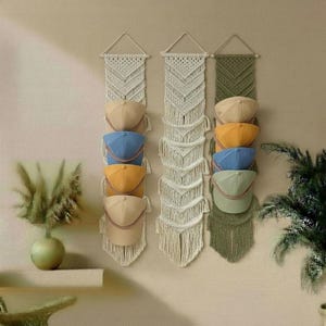 May include: Macrame hat organizers in cream and olive green, each holding baseball caps in various colors. The caps include tan, blue, yellow, and green. The organizers are hanging on a beige wall, with a vase and plant in the background.