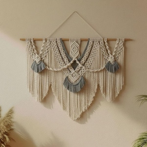 Può includere: Applique da parete in macramè bianco sporco con dettagli grigi. Il design presenta nodi intricati e nappe decorative, sospese a un tassello di legno. Un pezzo in stile bohémien, adatto all'arredamento d'interni.