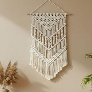 Op de afbeelding: Crèmekleurige macramé wandhanger met een geometrisch ontwerp. De wanddecoratie heeft een houten deuvel aan de bovenkant om op te hangen, met ingewikkelde knoop patronen en een gefranjerde onderkant. Dit stuk in bohemien stijl voegt textuur en warmte toe aan elke ruimte.