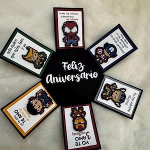 May include: A circular arrangement of greeting cards with pixelated superhero designs. The central card reads "Feliz Aniversario." Each card features a different character and a Spanish message, with various color borders.