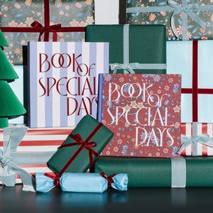 Puede incluir: Dos libros titulados Book of Special Days están rodeados de varios paquetes de regalo. Un libro tiene una cubierta a rayas azul claro y blancas, mientras que el otro presenta un patrón floral rojo. Los regalos están envueltos en papel verde sólido, a rayas rojas y floral con cintas de terciopelo.