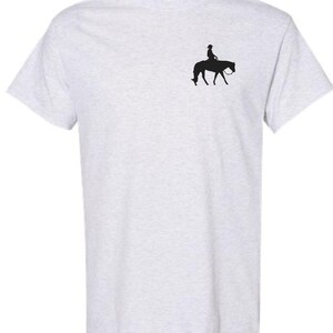 May include: Light gray t-shirt featuring a small black silhouette of a person riding a horse. The rider is wearing a hat. The horse is facing left, with its head lowered. The shirt is made of a soft material.