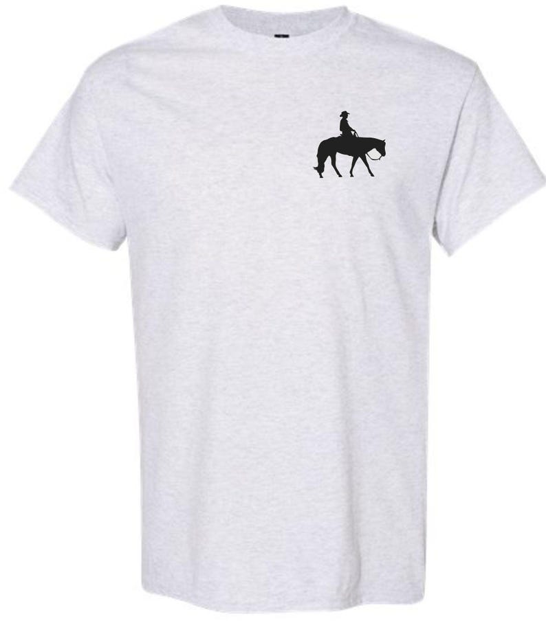 May include: Light gray t-shirt featuring a small black silhouette of a person riding a horse. The rider is wearing a hat. The horse is facing left, with its head lowered. The shirt is made of a soft material.