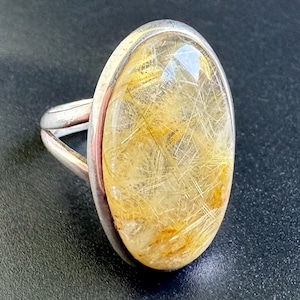 May include: A silver ring featuring a large, oval-shaped rutilated quartz gemstone. The stone is translucent with visible golden, needle-like inclusions throughout. The metal band is smooth and polished, securely holding the cabochon-cut stone.