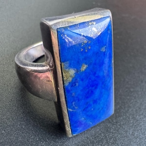 May include: A silver ring featuring a large, rectangular, deep blue lapis lazuli gemstone set in a smooth metal bezel. The stone displays natural golden pyrite inclusions across its surface, and the band is a solid, polished silver metal.