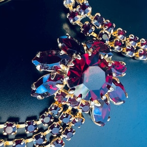 May include: A gold-tone metal necklace featuring a large central floral-shaped cluster of deep red faceted stones. The design includes multiple rows of smaller, square-cut red stones set in metal links that extend from the central floral piece.