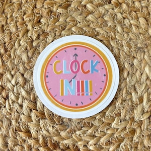 May include: A round sticker with a pink clock face design. The sticker has the words "CLOCK IN!!!" in colorful letters. The clock hands point upwards. The sticker has a white border and an orange and yellow outer ring.