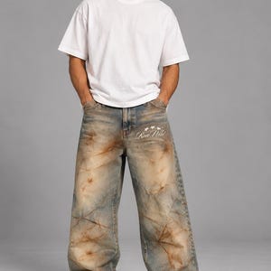 Könnte beinhalten: Ein schlichtes weißes Kurzarm-T-Shirt und eine weit geschnittene Jeans mit verwaschener blau-brauner Batik-Optik. Die Jeans ist im oberen vorderen Oberschenkelbereich mit dem weißen Sticktext Run Wild und drei Sternen darüber versehen.