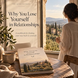 May include: A softcover workbook titled Why You Lose Yourself in Relationships by The Quiet Between. The cover features a landscape illustration of a villa among cypress trees. The book rests on a wooden table next to a ceramic mug.