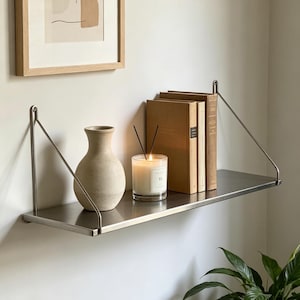May include: A stainless steel wall shelf with a vase, books, and a candle. The shelf is supported by angled metal brackets. The vase is beige, the books are brown, and the candle is in a glass jar.