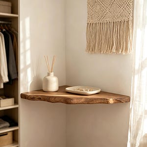 May include: A wooden corner shelf with a live edge. A small, off-white ceramic vase with reeds and a small, off-white ceramic dish with jewelry sit on the shelf. A woven wall hanging with fringe is above the shelf.