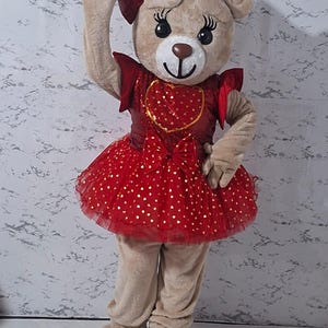 Könnte beinhalten: Ein Ganzkörper-Teddybär-Kostüm in Beige und Rot. Der Bär hat eine rote Schleife auf dem Kopf und trägt ein rotes Kleid mit goldenen Herzakzenten. Das Kostüm beinhaltet Handschuhe und große Füße.