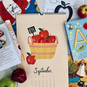 May include: A calendar page for September features a watercolor illustration of a basket filled with red apples. The basket is yellow with pink stripes. The text "September" is written in cursive. Books and apples surround the calendar.