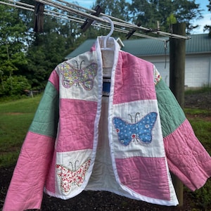 May include: A quilted jacket featuring a patchwork design of pink and white squares with green sleeves. The front panels are decorated with three distinct butterfly appliqués in floral, blue, and red patterns, finished with a white trim border.
