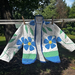 May include: A quilted jacket featuring a white base with blue flower appliqués, yellow centers, and green leaf accents. The garment has long sleeves, a blue trim along the open front, and a horizontal green and blue band at the bottom hem.