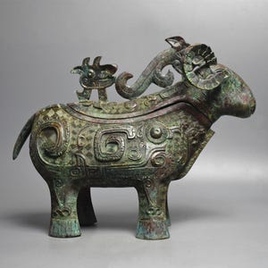 Puede incluir: Escultura de carnero de bronce con intrincados patrones y una pequeña figura con forma de dragón en su parte posterior. La escultura tiene un acabado de pátina, con tonos verdes y marrones. El carnero tiene cuernos grandes y curvados.