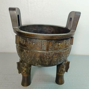 May include: A bronze-colored, antique-style vessel with two handles and three decorative legs. The vessel features intricate patterns around the body and is likely used for decorative or ceremonial purposes. The overall design suggests a historical or cultural significance.