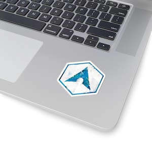 Peut inclure: Un autocollant blanc avec un motif géométrique bleu et blanc, représentant une flèche stylisée dans un cadre hexagonal. L'autocollant est placé sur un ordinateur portable argenté, avec le clavier visible.
