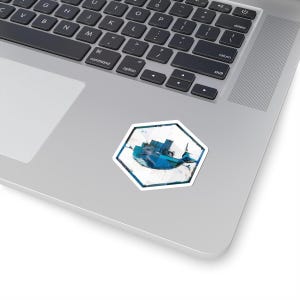 May include: A white, hexagonal sticker featuring a blue abstract design. The design appears to be a stylized depiction of a whale or a similar aquatic creature. The sticker is placed on a silver laptop.