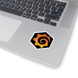 Puede incluir: Una pegatina hexagonal con un diseño abstracto. La obra de arte presenta una espiral central en tonos naranja y amarillo, sobre un fondo negro. La pegatina tiene un borde blanco y está colocada en un portátil.