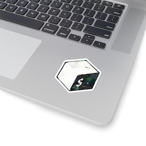 Könnte beinhalten: Ein weißer, schwarzer und grüner geometrischer Aufkleber in sechseckiger Form. Der Aufkleber befindet sich auf einem silbernen Laptop. Der Aufkleber hat einen weißen Rand und ein Design, das eine Kombination aus abstrakten Formen und Farben zu sein scheint.