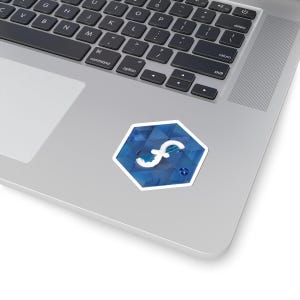 Op de afbeelding: Een blauw-witte zeshoekige sticker met een wit gestileerd "f"-symbool. De sticker heeft een textuur die op een edelsteen lijkt en is geplaatst op een zilveren laptop.